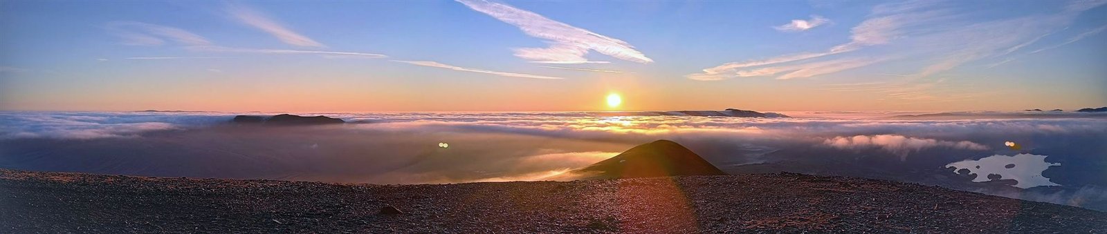 Panorama of sunrise over a sea of cloud filling the valleys, seen from a stony summit ridge