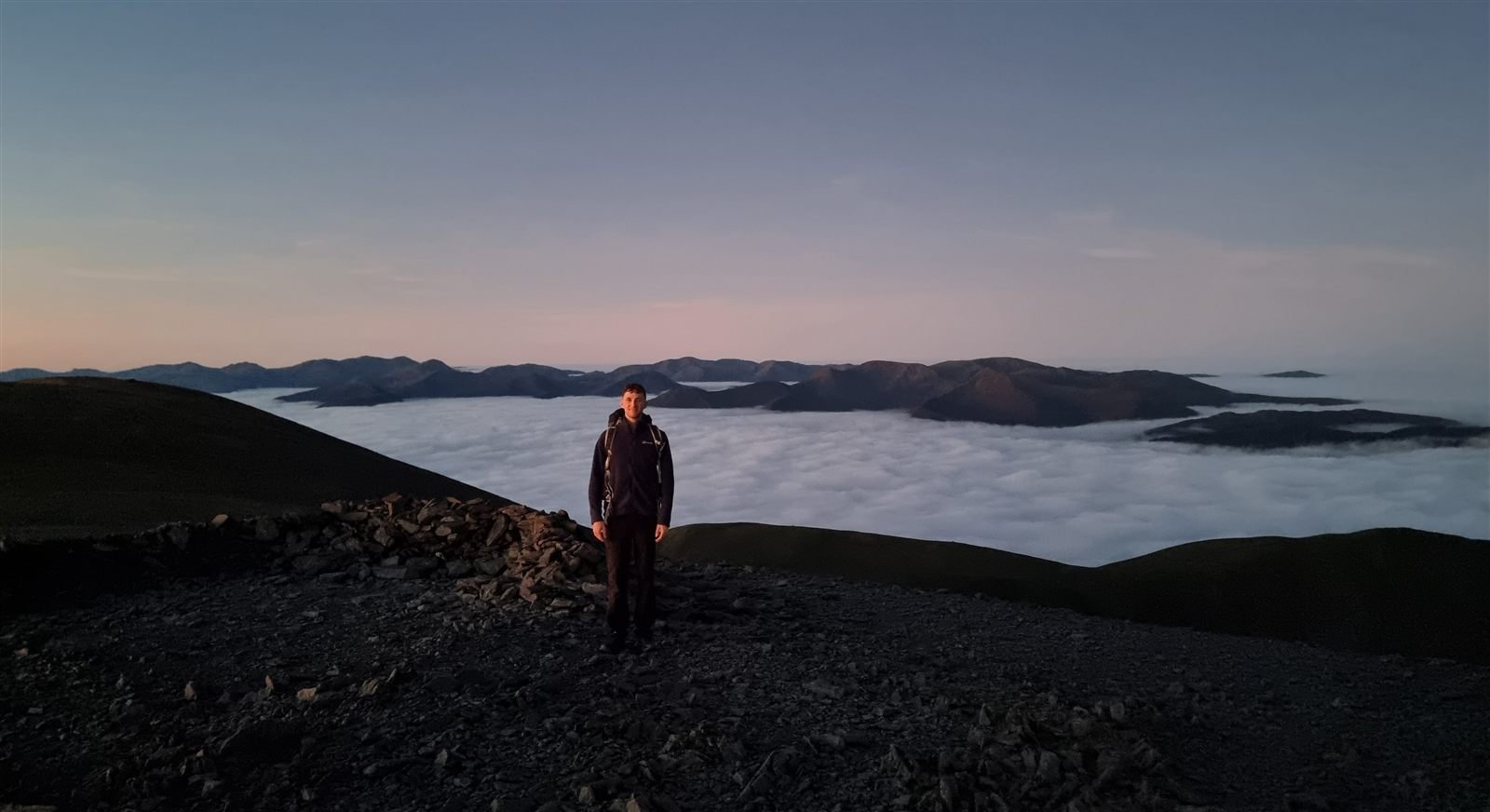 Joss standing on a stony summit above a sea of cloud at dawn, with fells rising through the cloud behind