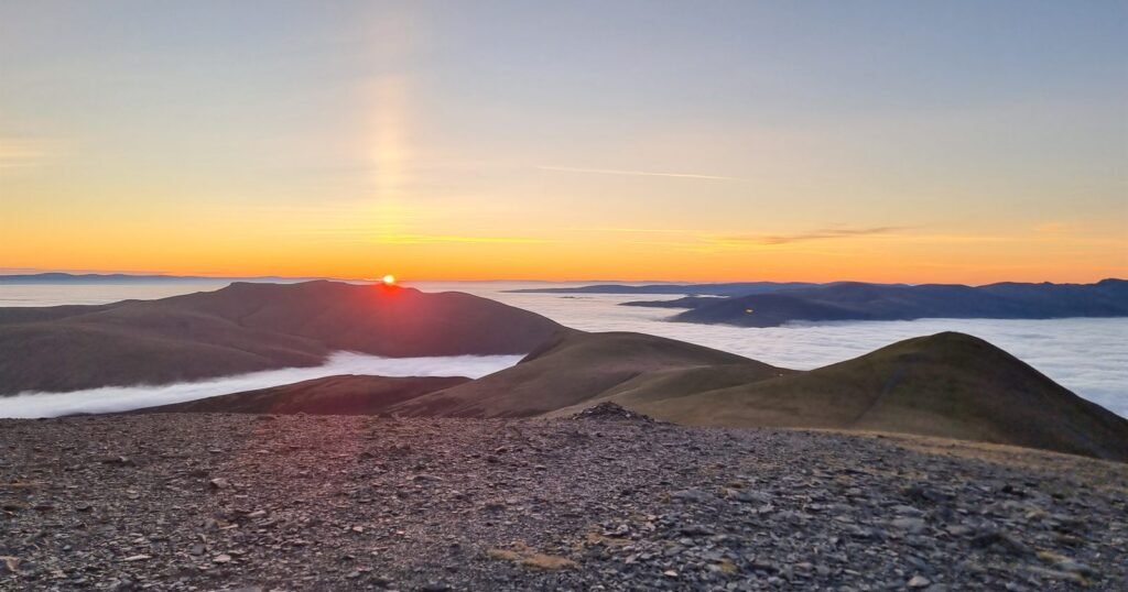 The sun rising over a fell seen from the stony summit of Skiddaw, with cloud filling the valleys below under an orange dawn sky.