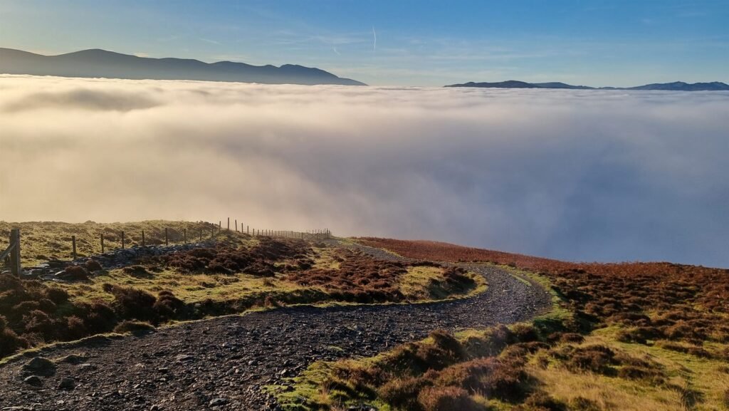 A stony track curving through heather and bracken on Skiddaw to the edge of a sunlit cloud sea, with fells on the horizon under a blue sky.