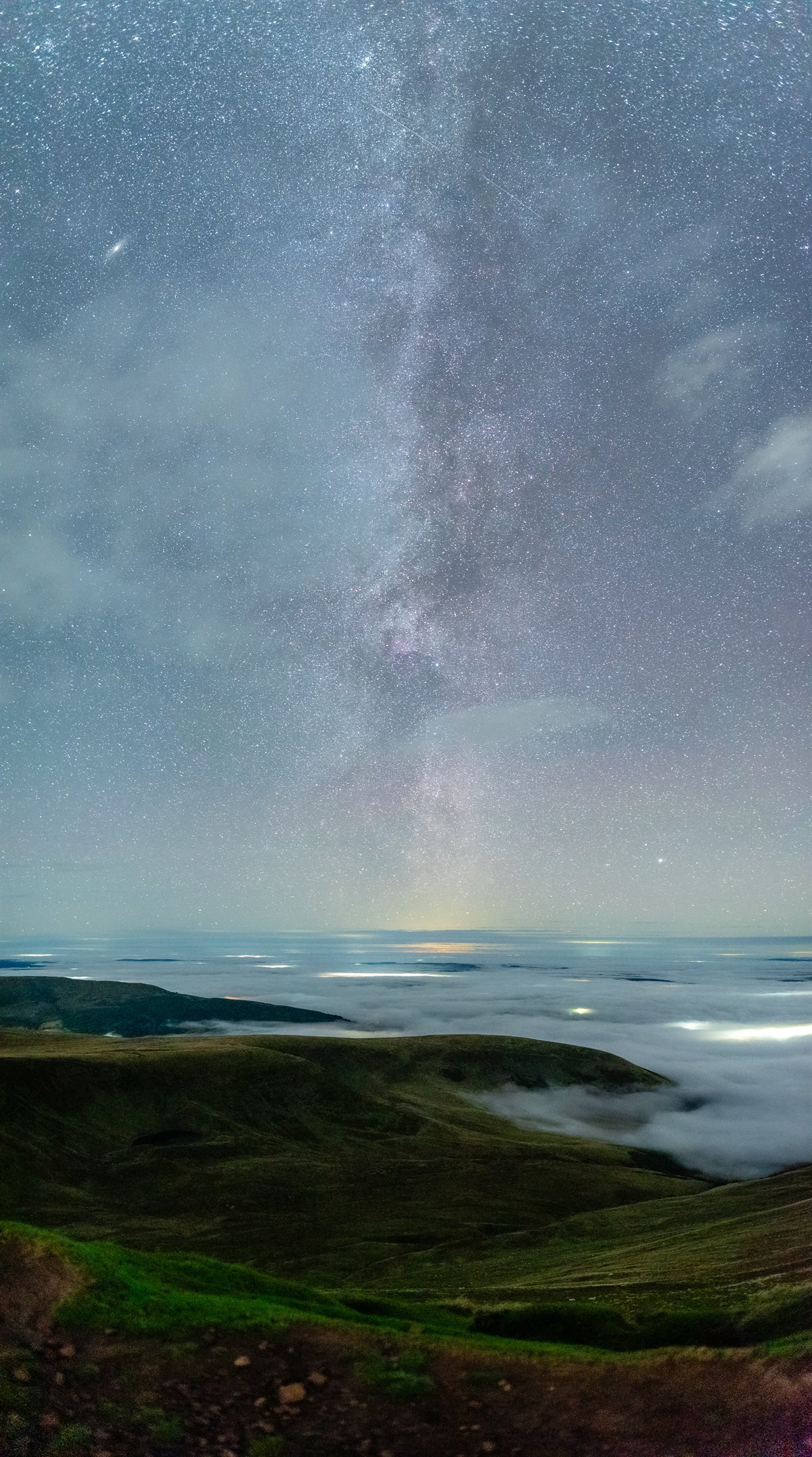 Pen y Fan before dawn on 22 September 2026: the Milky Way above a sea of mist filling the valleys below the grassy Bannau Brycheiniog ridges, with town lights glowing through the cloud.