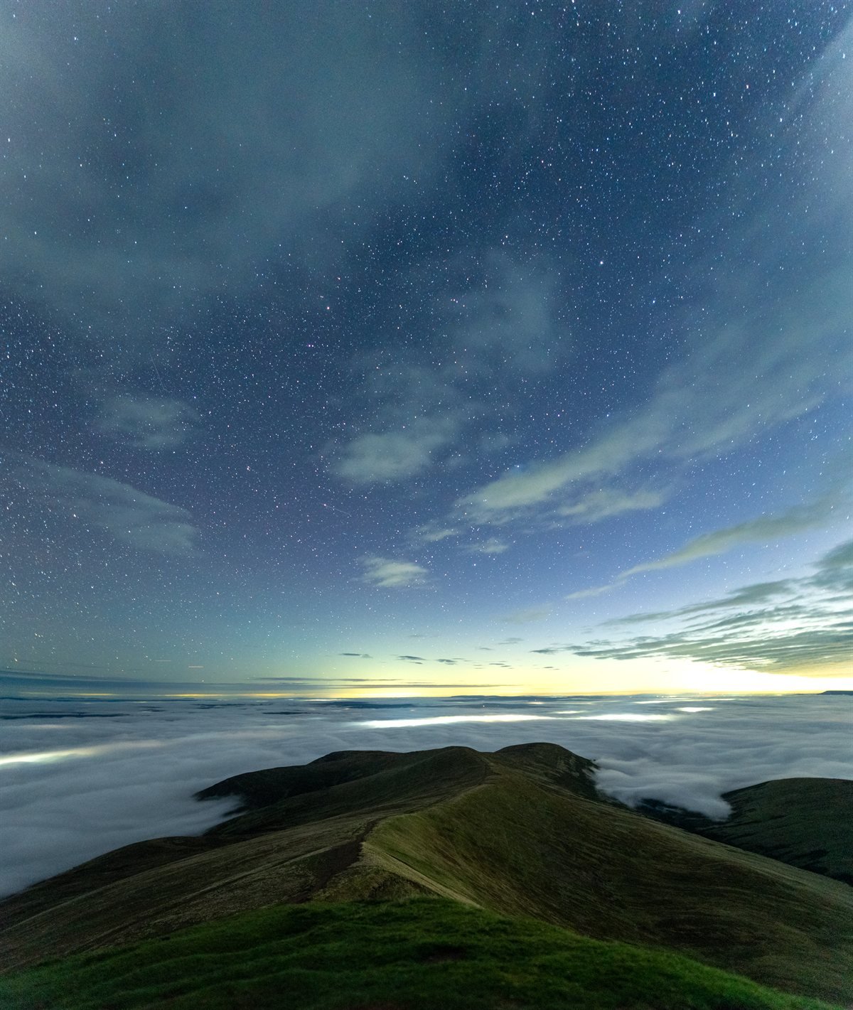 A grassy ridge path in Bannau Brycheiniog before dawn on 22 September 2026, with a cloud sea filling the valleys and the first light glowing on the horizon under a starry sky.