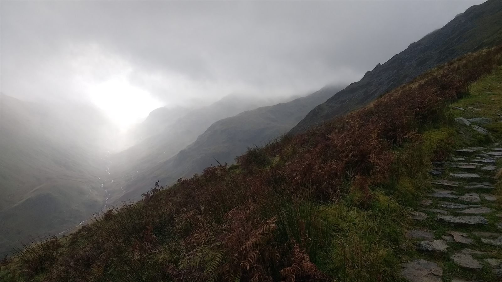 A pitched stone path climbing a bracken covered fellside beside a deep valley filled with grey mist and low cloud, with a pale sun glowing through the murk.