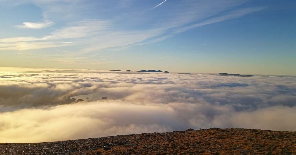 A bright white cloud sea filling the valleys under a blue sky with wispy high cloud, a few dark fell tops on the horizon, a lake with small islands showing through a gap in the cloud, and golden grass along the fellside in the foreground.