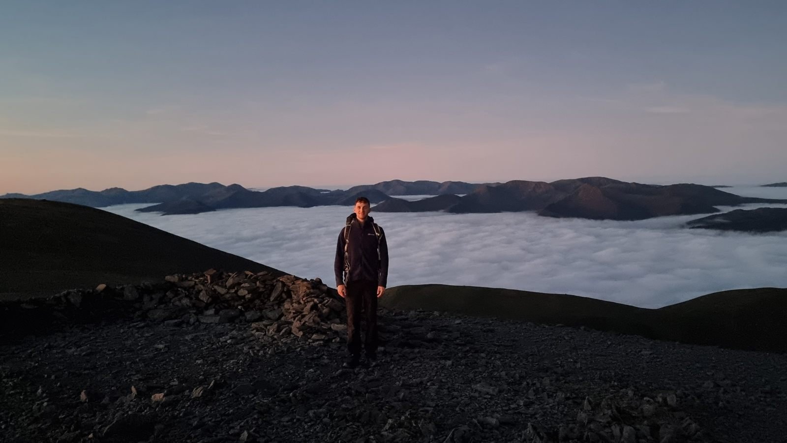 Joss standing by the stone shelter on the summit of Skiddaw before sunrise, with a cloud sea and a chain of fells behind him.
