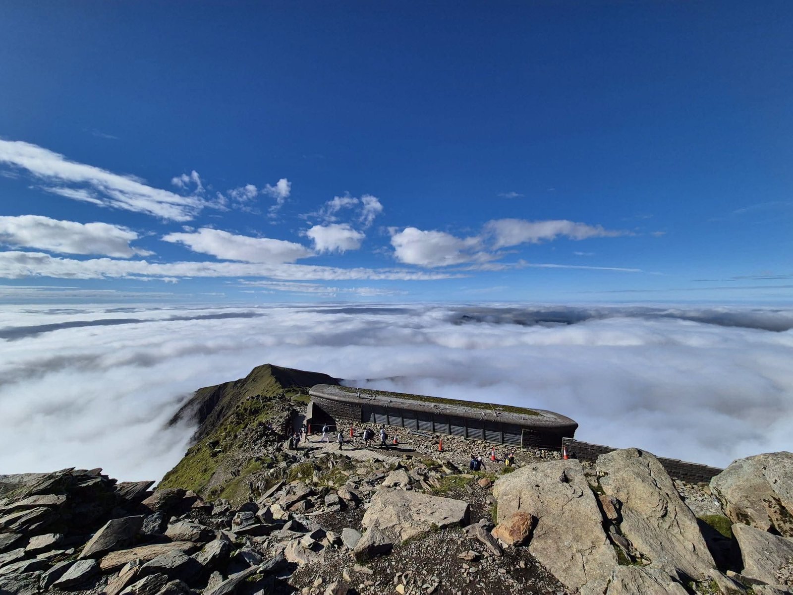 The summit of Yr Wyddfa on 22 September 2026: the Hafod Eryri building and walkers in bright sun above an unbroken cloud sea filling every valley, with only the summit ridge and one distant top breaking through.
