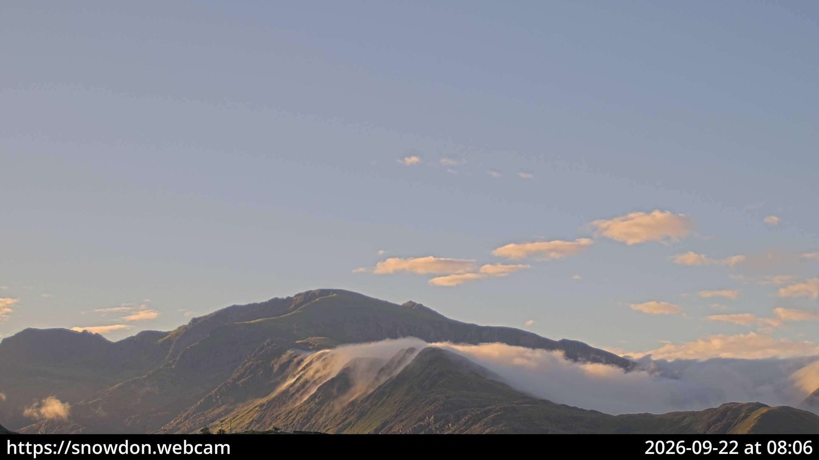 Webcam frame at 08:06 BST on 22 September 2026: Yr Wyddfa clear against blue sky in golden light, with cloud pouring over a lower ridge below the summit and a cloud sea to the right.